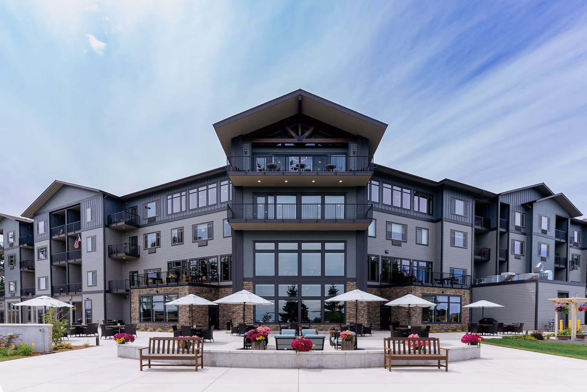 Front exterior view of a modern multi-story retirement community building with balconies, large windows, outdoor seating areas with umbrellas, benches, and flower pots under a partly cloudy sky.
