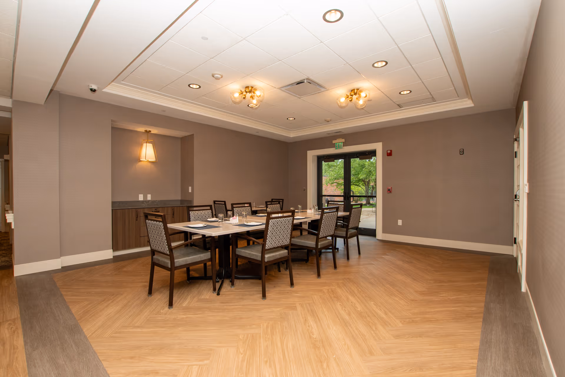 A dining room with a rectangular table set for six people, surrounded by six chairs with patterned upholstery. The room has light brown walls, a wooden floor with a darker border, ceiling lights, and a glass door leading outside with greenery visible through it.