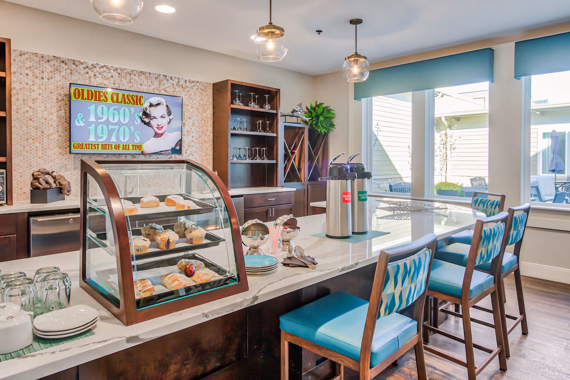 A bright dining area with a marble countertop island featuring a glass display case with pastries, coffee dispensers, plates, and utensils. There are four high chairs with blue patterned cushions around the island. Behind the counter, there are dark wooden cabinets with glassware and a wine rack. A colorful retro-style sign on the wall reads 'OLDIES CLASSIC & 1960's 1970's GREATEST HITS OF ALL TIME'. Large windows let in natural light and show an outdoor patio area.