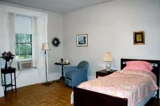 A simple bedroom with a pink quilted bed, nightstand, chair, lamp, and window.