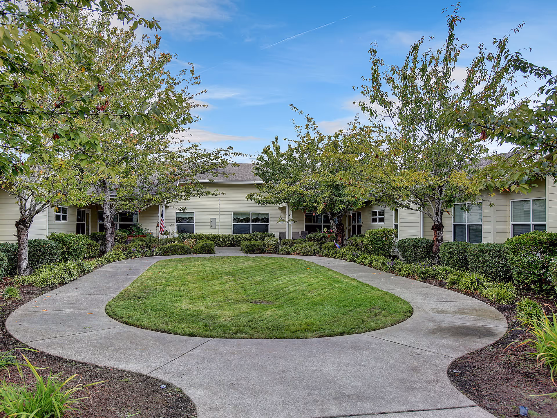 A landscaped outdoor courtyard area with a circular concrete walkway surrounding a green grassy patch. The courtyard is bordered by small trees and shrubs, with a single-story beige building in the background under a partly cloudy blue sky.