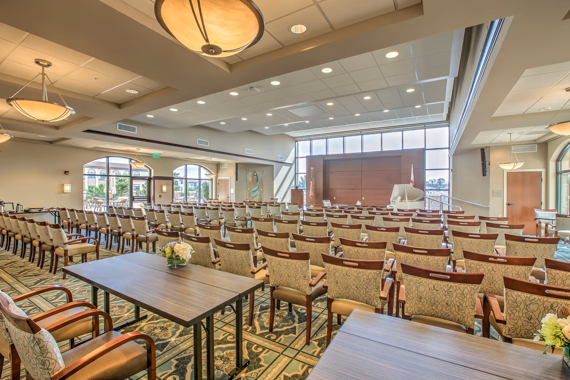 A spacious, well-lit conference or meeting room with rows of upholstered chairs arranged facing a stage area with a podium and a white grand piano. The room features large windows allowing natural light, decorative ceiling lights, and patterned carpet flooring. There are tables with flower arrangements in the foreground.