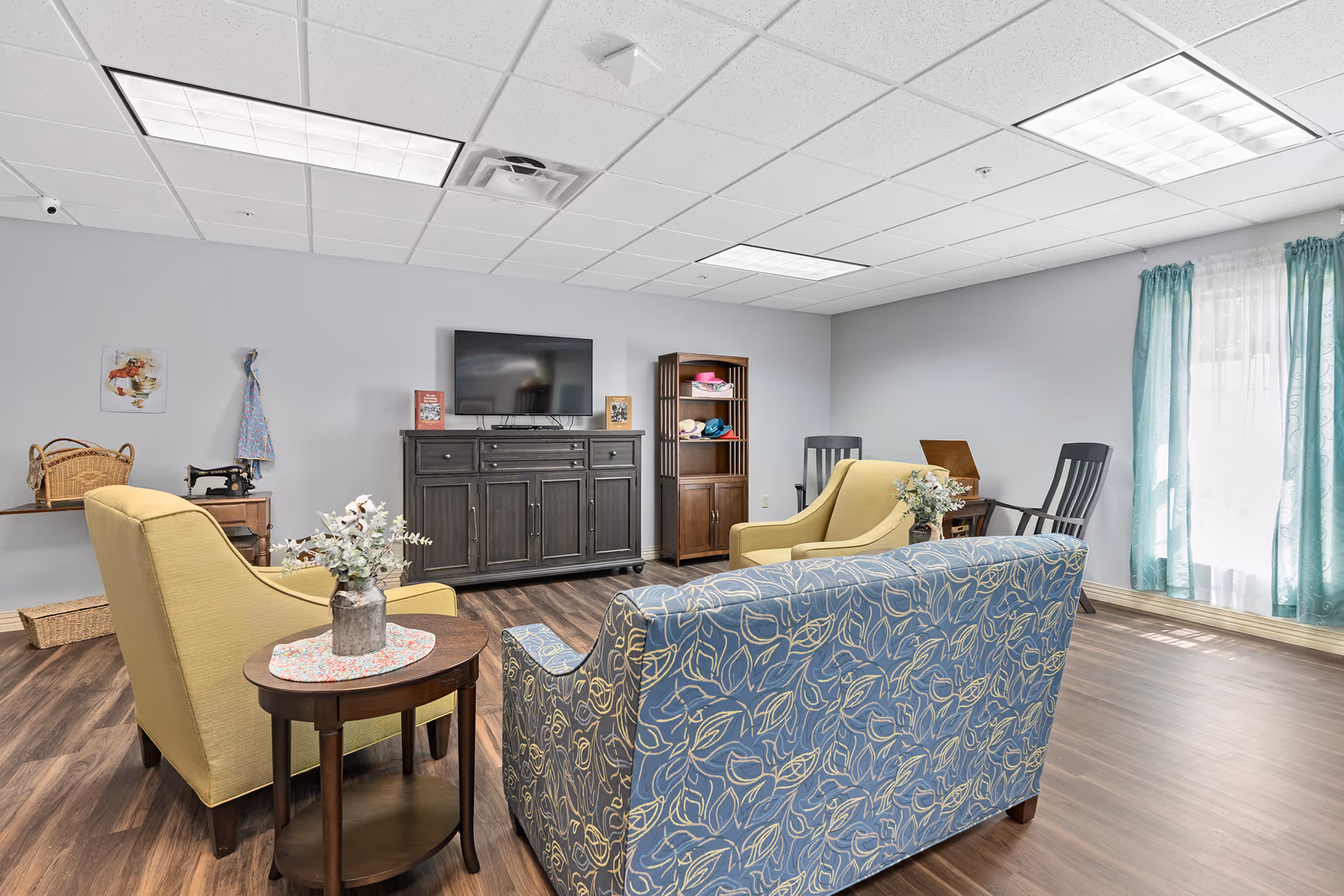 Bright common living room with sofas and armchairs arranged around a TV cabinet, wood-look flooring and a window with teal curtains.