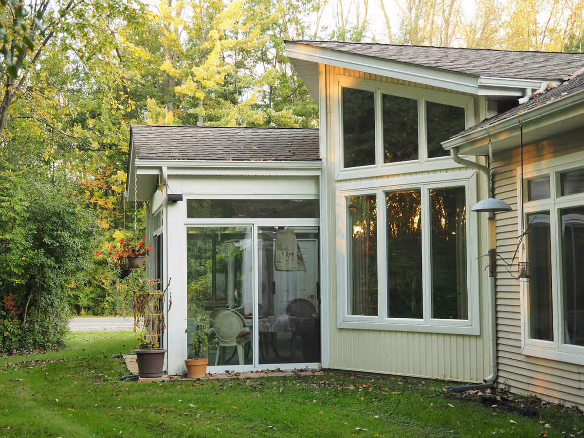Exterior view of a residential building with large windows and a sliding glass door leading to a small patio area. The building is surrounded by green grass and trees with some autumn foliage. There are potted plants near the patio and a hanging light fixture on the side of the building.