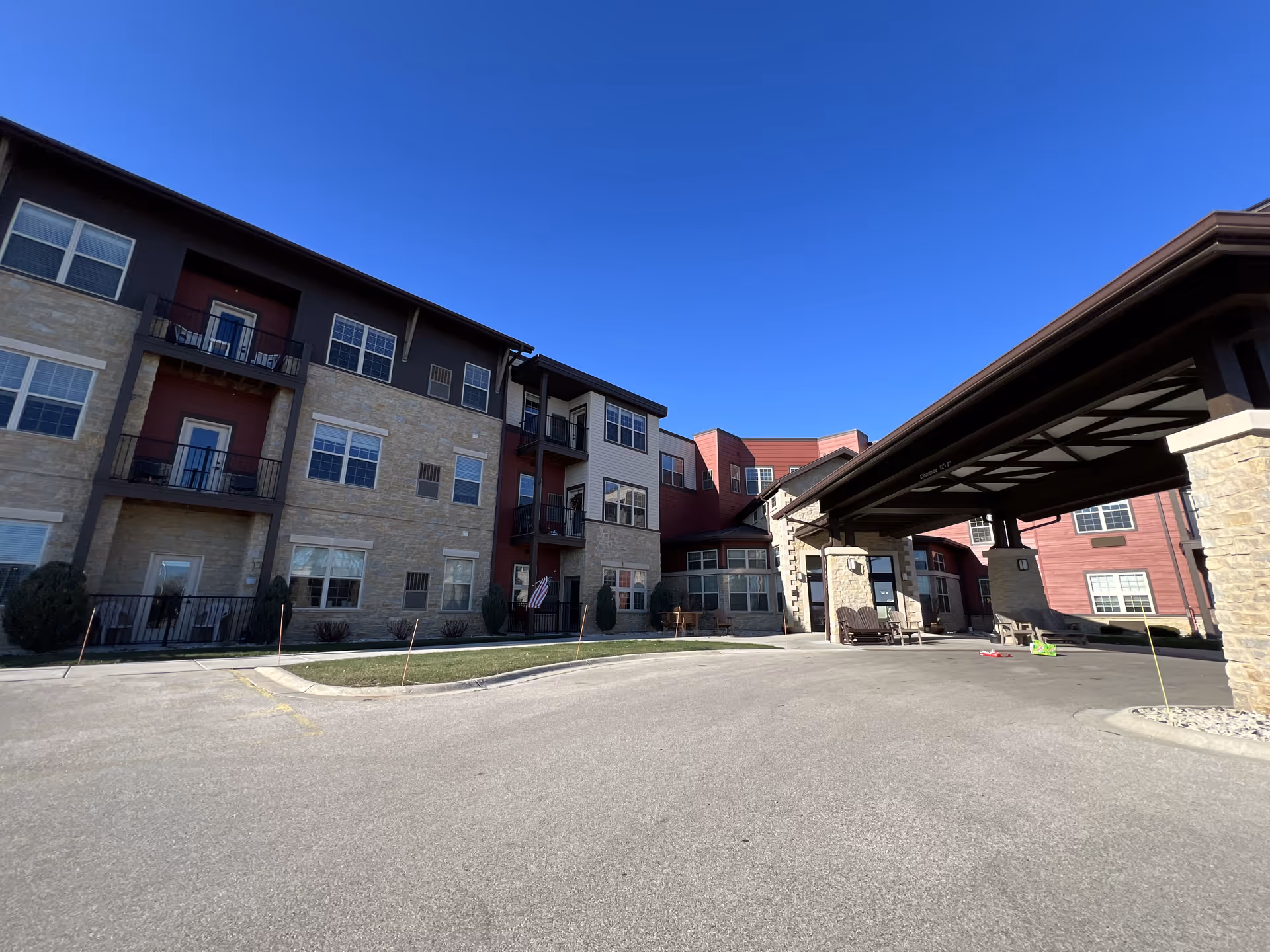 Exterior view of a senior living facility building with multiple floors, balconies, and a covered entrance area under a clear blue sky.
