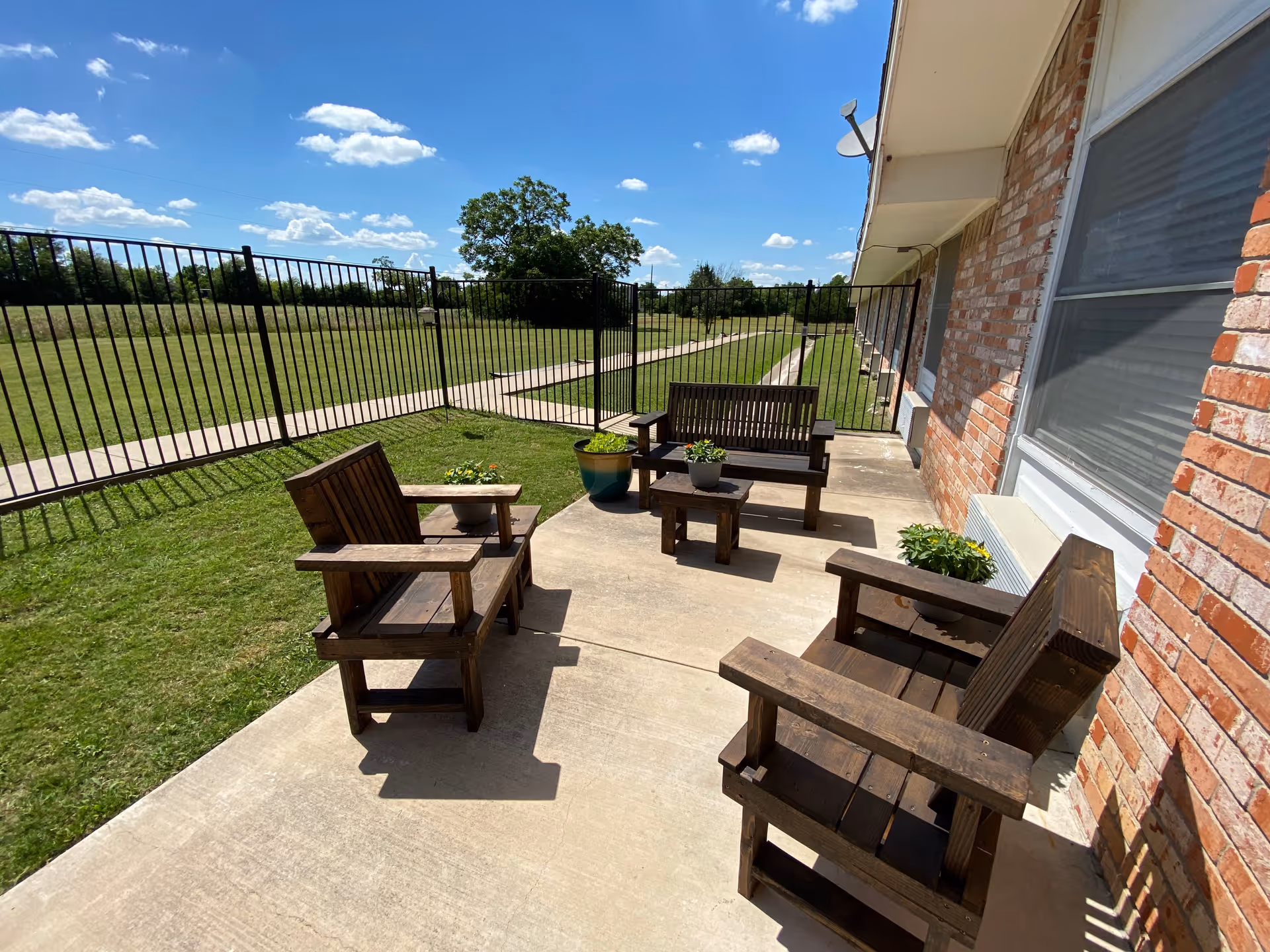 Outdoor patio with wooden chairs and a bench on a concrete walkway beside a brick building overlooking a fenced grassy yard under a blue sky.