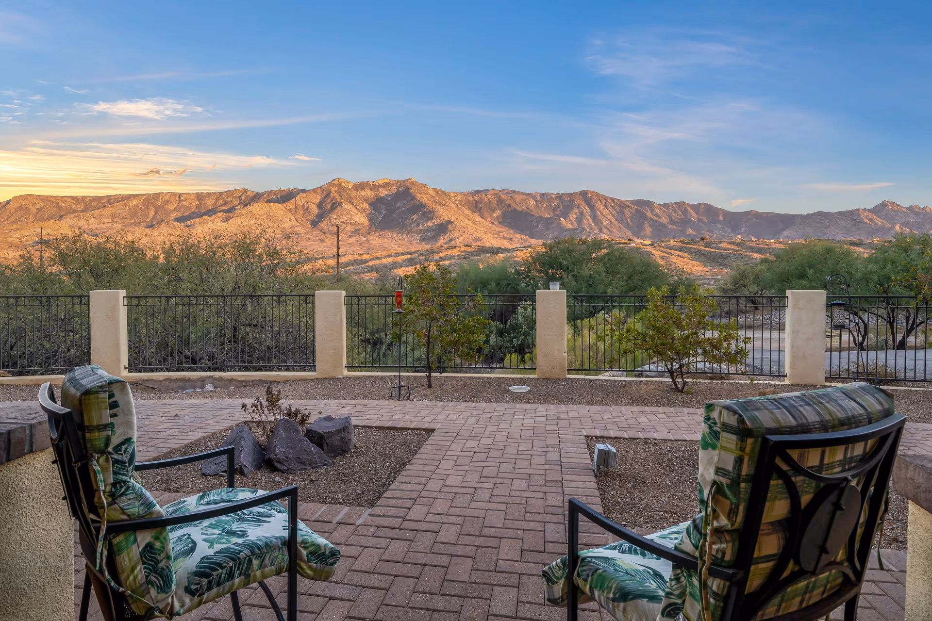 A patio with two cushioned chairs overlooking a fenced desert landscape and distant mountains at sunset.