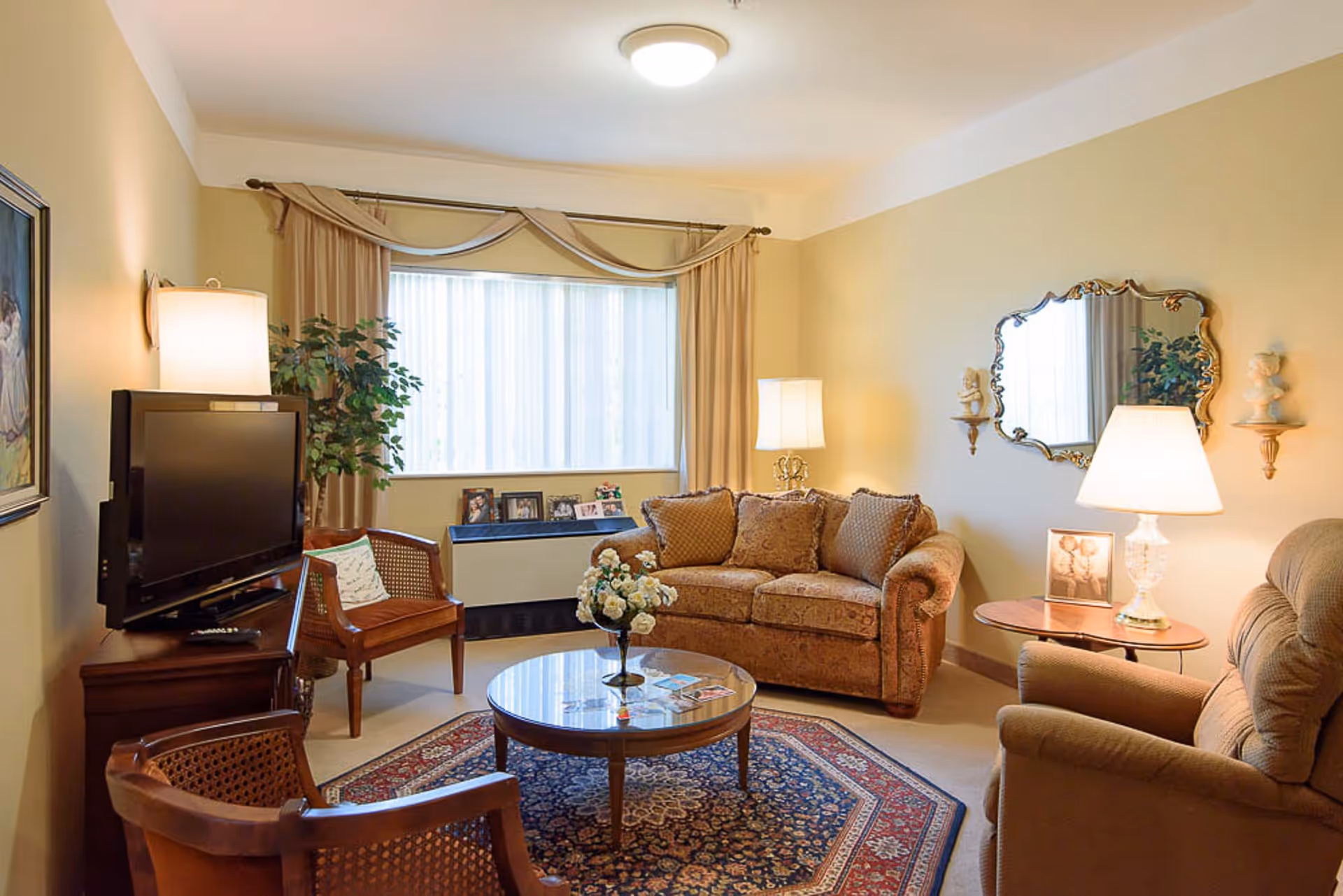 A cozy living room with a patterned sofa, an armchair, two wooden chairs, a round glass coffee table with a flower vase, a TV on a wooden stand, a large window with beige curtains, a decorative mirror on the wall, and two table lamps providing warm lighting.