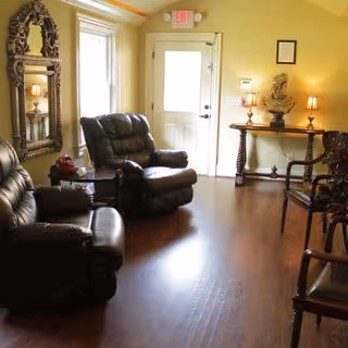 Small sitting room with leather recliners, wooden chairs, a console table with lamps and a decorative mirror near an exterior door.