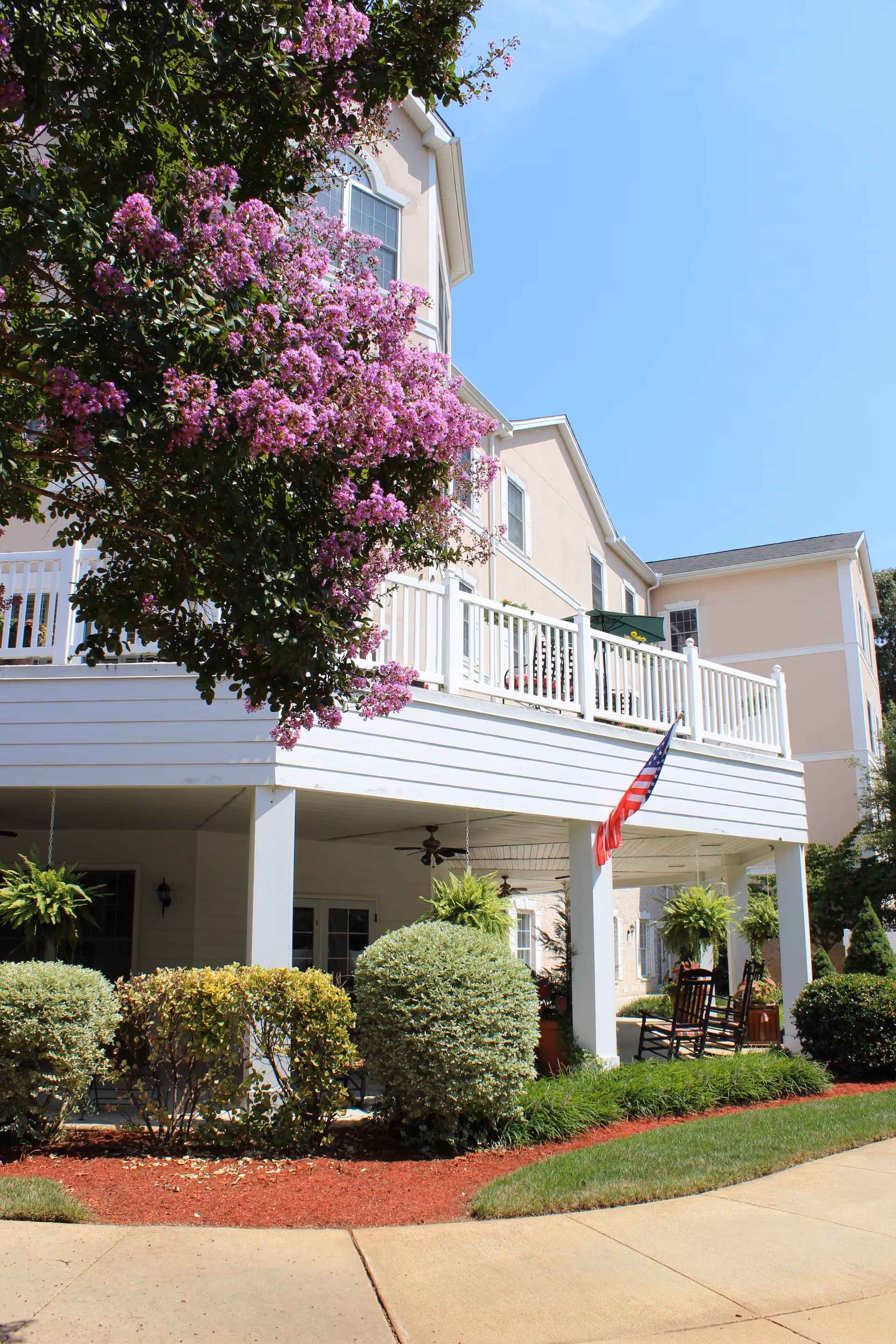 Exterior view of a senior living facility building with beige walls and white railings. There is a porch area with rocking chairs, hanging plants, and an American flag. In the foreground, there are well-maintained bushes, a flowering tree with purple blossoms, and a paved walkway under a clear blue sky.