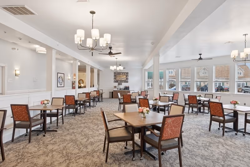 A spacious dining room with multiple square wooden tables, each surrounded by four chairs with patterned orange and beige upholstery. The room has large windows letting in natural light, beige carpet flooring, white walls, and ceiling lights with chandeliers and ceiling fans. Small floral centerpieces are placed on each table.