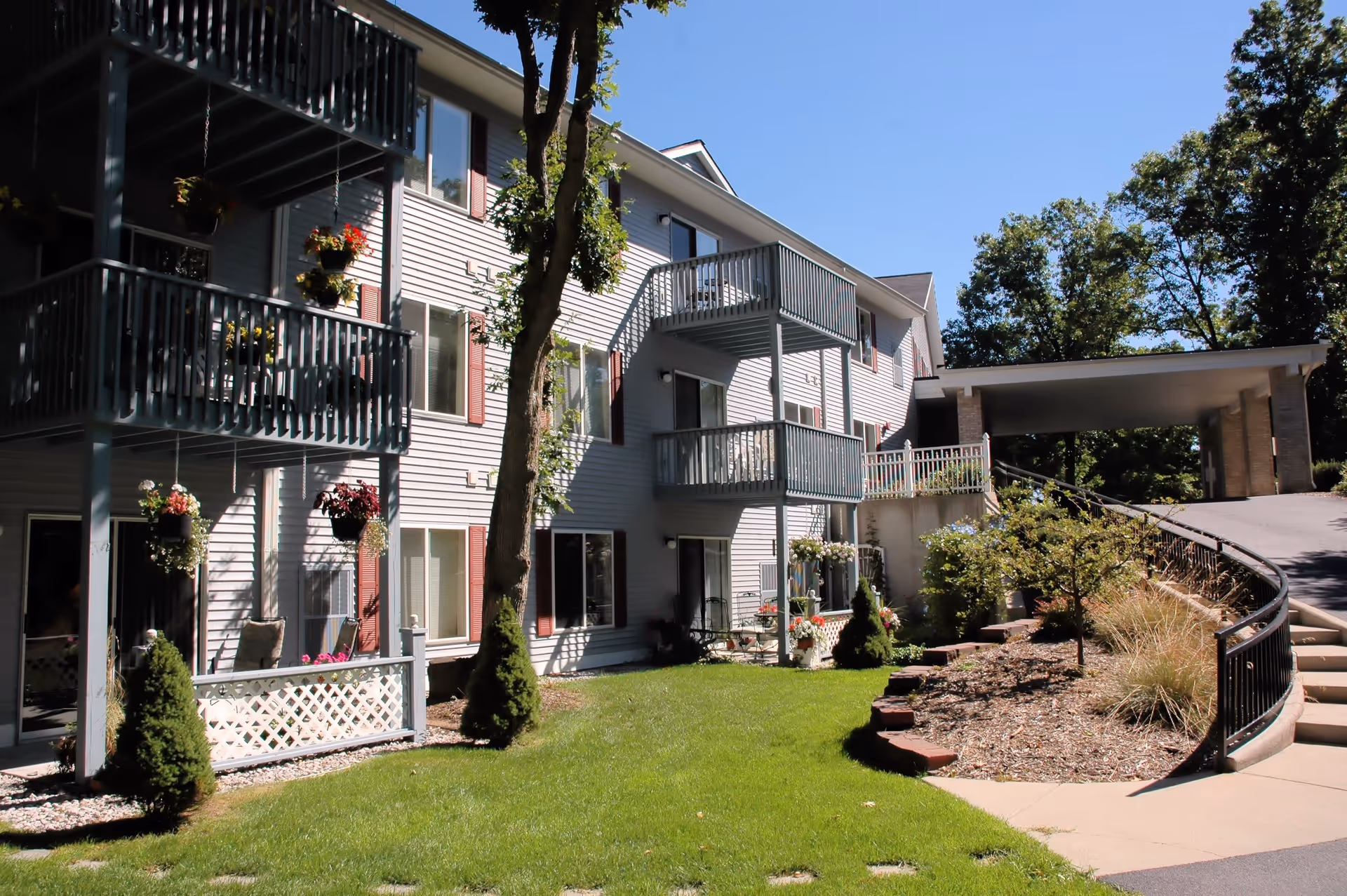 Exterior view of a multi-story residential building with balconies, hanging flower pots, and a well-maintained lawn and garden area under a clear blue sky.