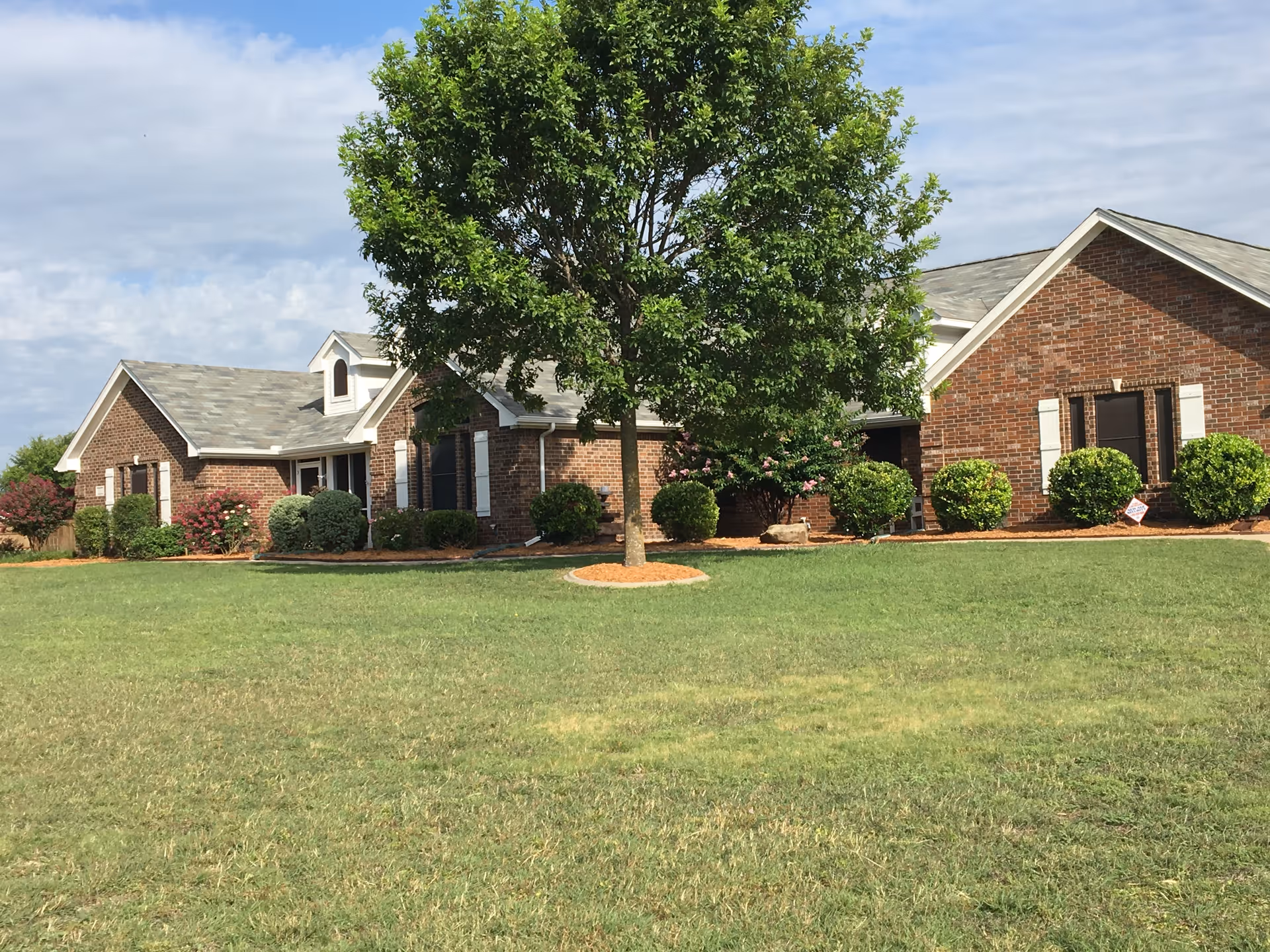 A single-story brick building with a gray shingled roof surrounded by green grass and landscaped bushes. A large tree stands prominently in the front yard under a partly cloudy sky.