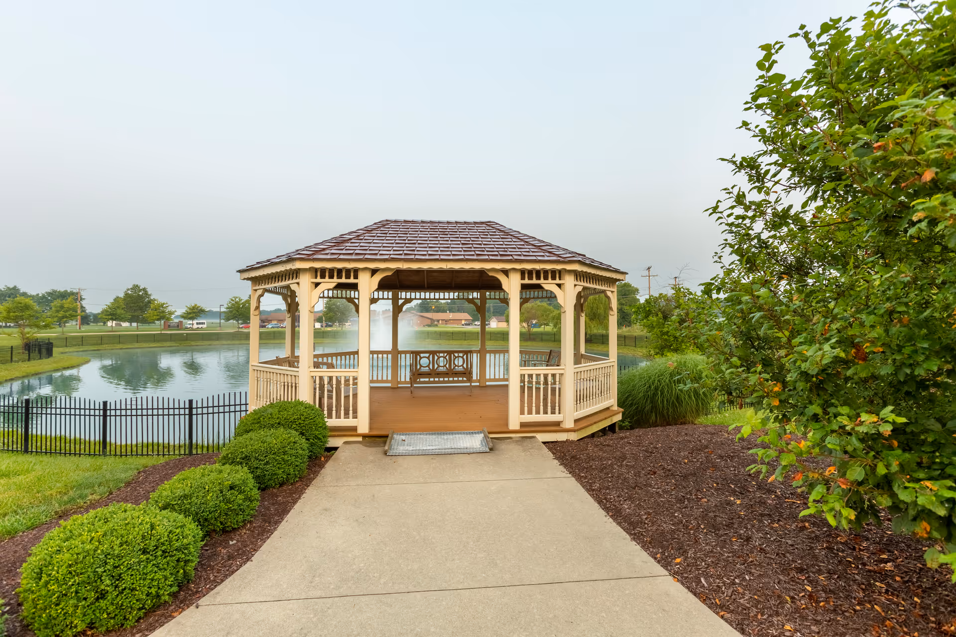 A wooden gazebo with a shingled roof situated at the end of a concrete pathway, surrounded by neatly trimmed bushes and greenery. In the background, there is a pond with a fountain and a black metal fence enclosing the area.