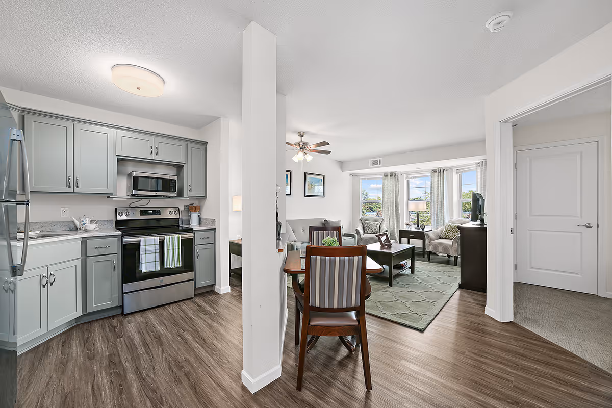 Interior view of a senior living apartment at The Rivers Retirement Community showing a modern kitchen with gray cabinets, stainless steel appliances, and a dining area with a wooden table and chairs. Beyond the dining area is a living room with a sofa, armchair, coffee table, and large windows with curtains letting in natural light. A ceiling fan is visible above the living room.