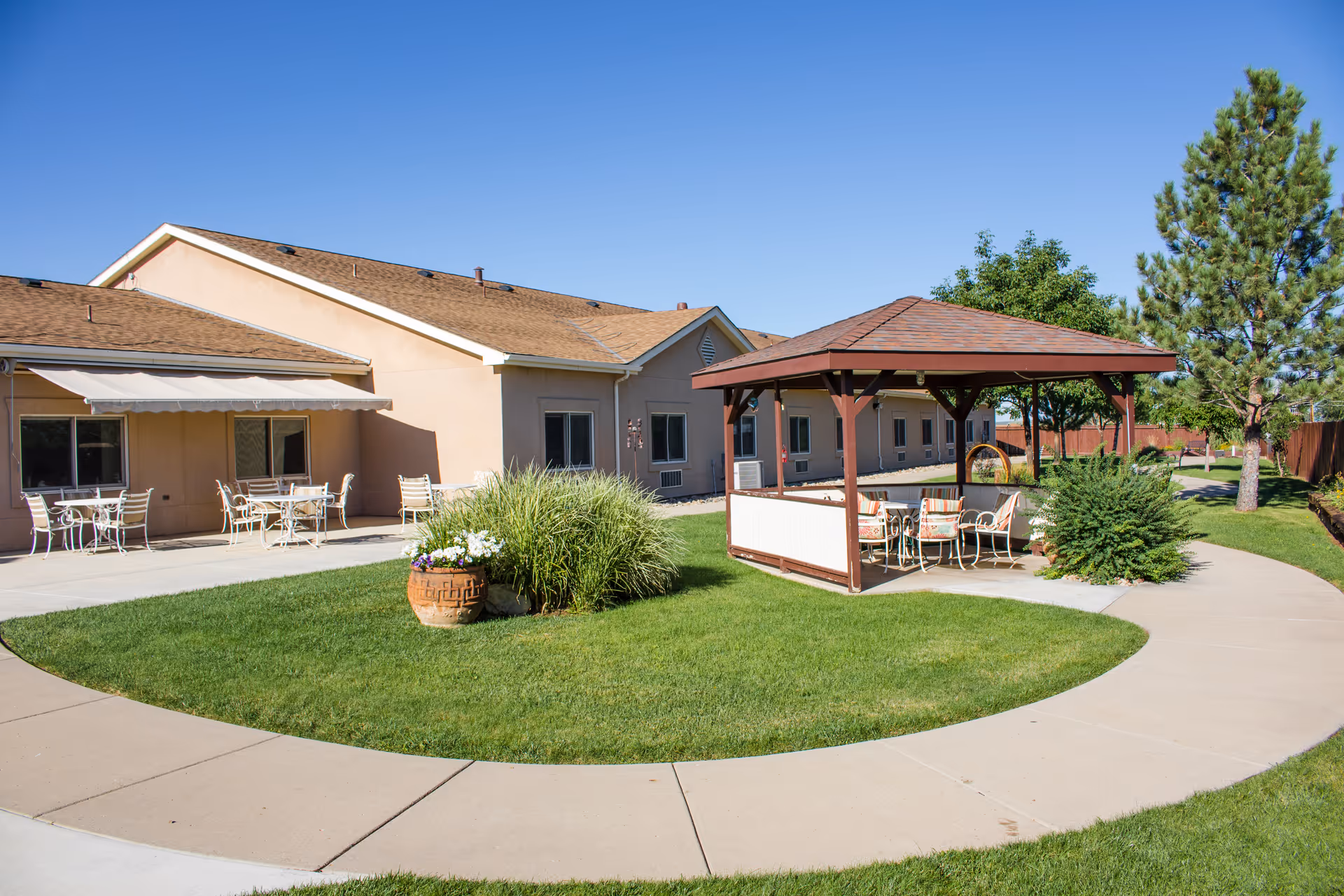 Outdoor area of Vista Mesa Assisted Living featuring a circular concrete walkway surrounding a grassy lawn with a wooden gazebo furnished with chairs and a table. The building exterior is beige with a brown roof, and there are additional tables and chairs on a patio area. Trees and shrubs are visible under a clear blue sky.