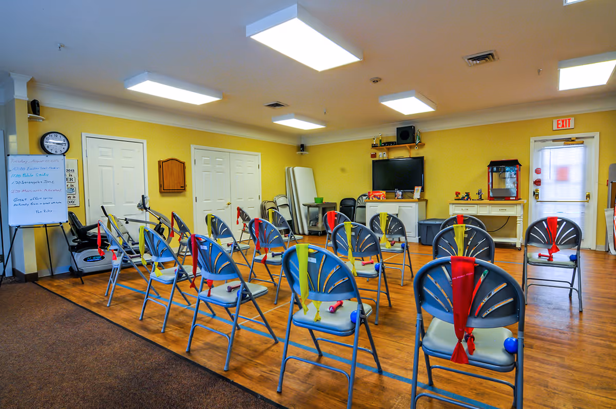A brightly lit room with rows of metal chairs arranged facing a television mounted on the wall. Each chair has colorful resistance bands and small dumbbells placed on or tied to them. The room has yellow walls, wooden flooring, and fluorescent ceiling lights. There is a whiteboard with a schedule written on it, a stationary exercise bike, and a popcorn machine near the exit door.