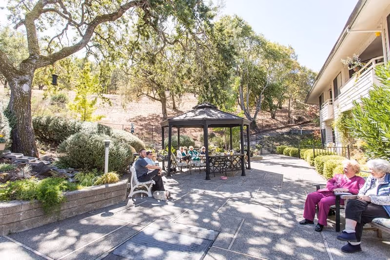 Outdoor patio area at Deer Park Retirement Community with several elderly people sitting on benches and chairs under a gazebo and around the patio. The area is surrounded by trees, bushes, and a hillside, with a building visible on the right side.