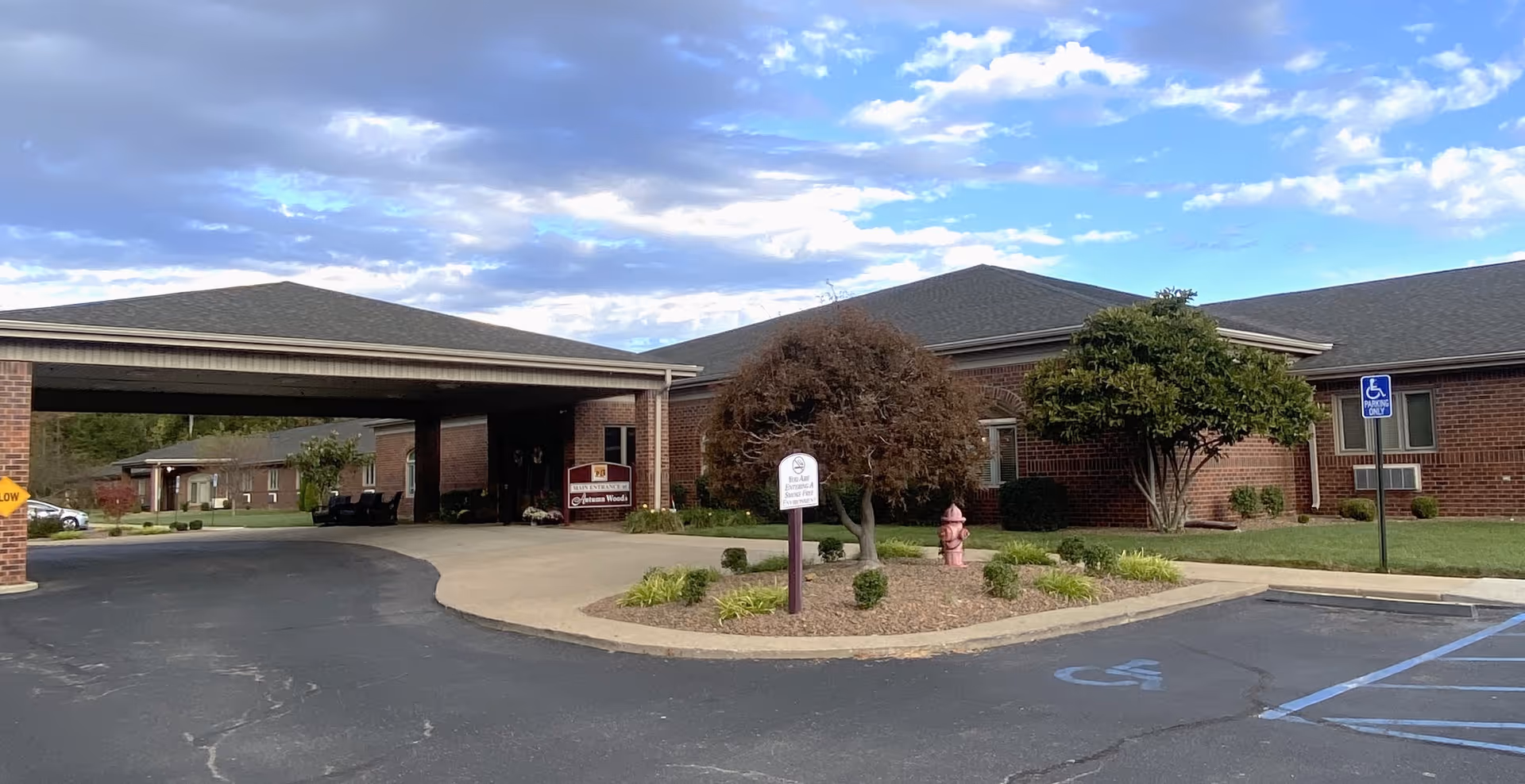 Exterior view of Autumn Woods Health Campus showing a brick building with a covered entrance driveway, landscaped area with trees and shrubs, a fire hydrant, and handicap parking spaces under a partly cloudy sky.