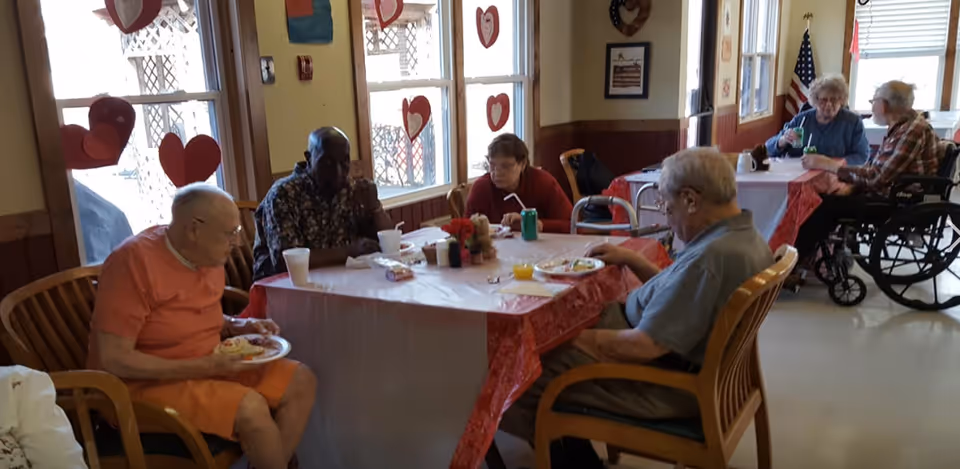 A group of elderly people sitting around two tables in a dining area decorated with heart-shaped cutouts on the windows. They are eating and drinking, with some using wheelchairs and others seated on wooden chairs. The room has large windows letting in natural light and an American flag in the background.