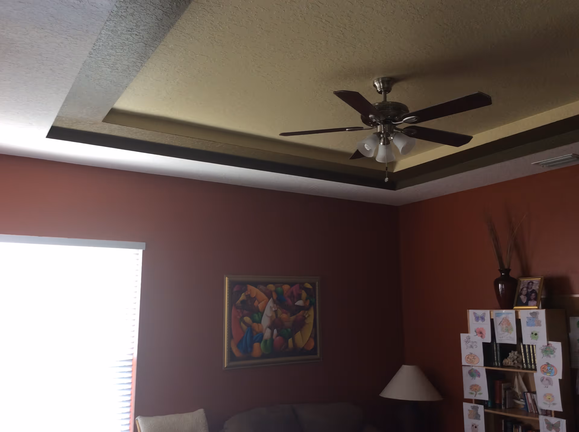 Interior view of a room with a textured ceiling featuring a ceiling fan with lights. The walls are painted dark red, and there is a window with blinds partially open letting in light. A colorful abstract painting is hung on the wall above a couch. To the right, there is a bookshelf decorated with various drawings and a vase with tall dried plants on top. A table lamp is also visible next to the bookshelf.