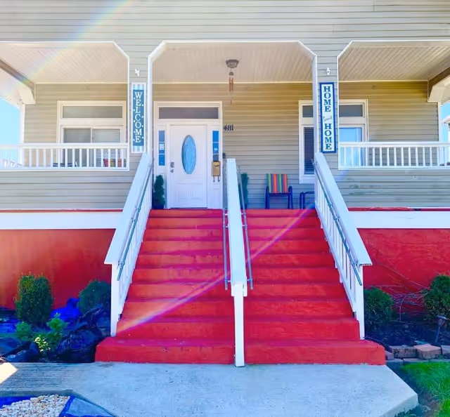 Front entrance of Montane Manor Assisted Living featuring a set of red stairs with white railings leading up to a white door with an oval window. The porch has two blue signs on either side of the door reading 'WELCOME' and 'HOME HOME'. There is a chair with a colorful striped cushion on the right side of the porch and some greenery around the base of the stairs.