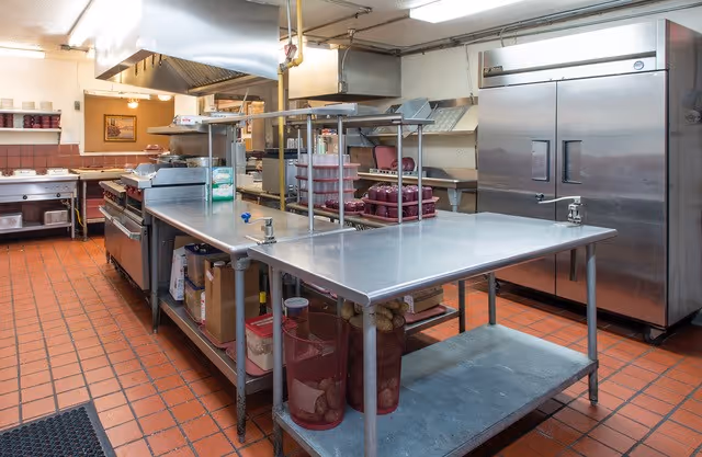 A commercial kitchen with stainless steel work tables, large refrigerators, an industrial stove, and various kitchen supplies. The floor is covered with red tiles and the walls are light-colored. The kitchen appears clean and well-organized.