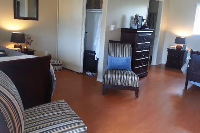 A senior living facility bedroom with wooden flooring, two striped chairs with blue cushions, a dark wooden dresser, two nightstands with lamps, and an open closet with a white robe hanging inside.
