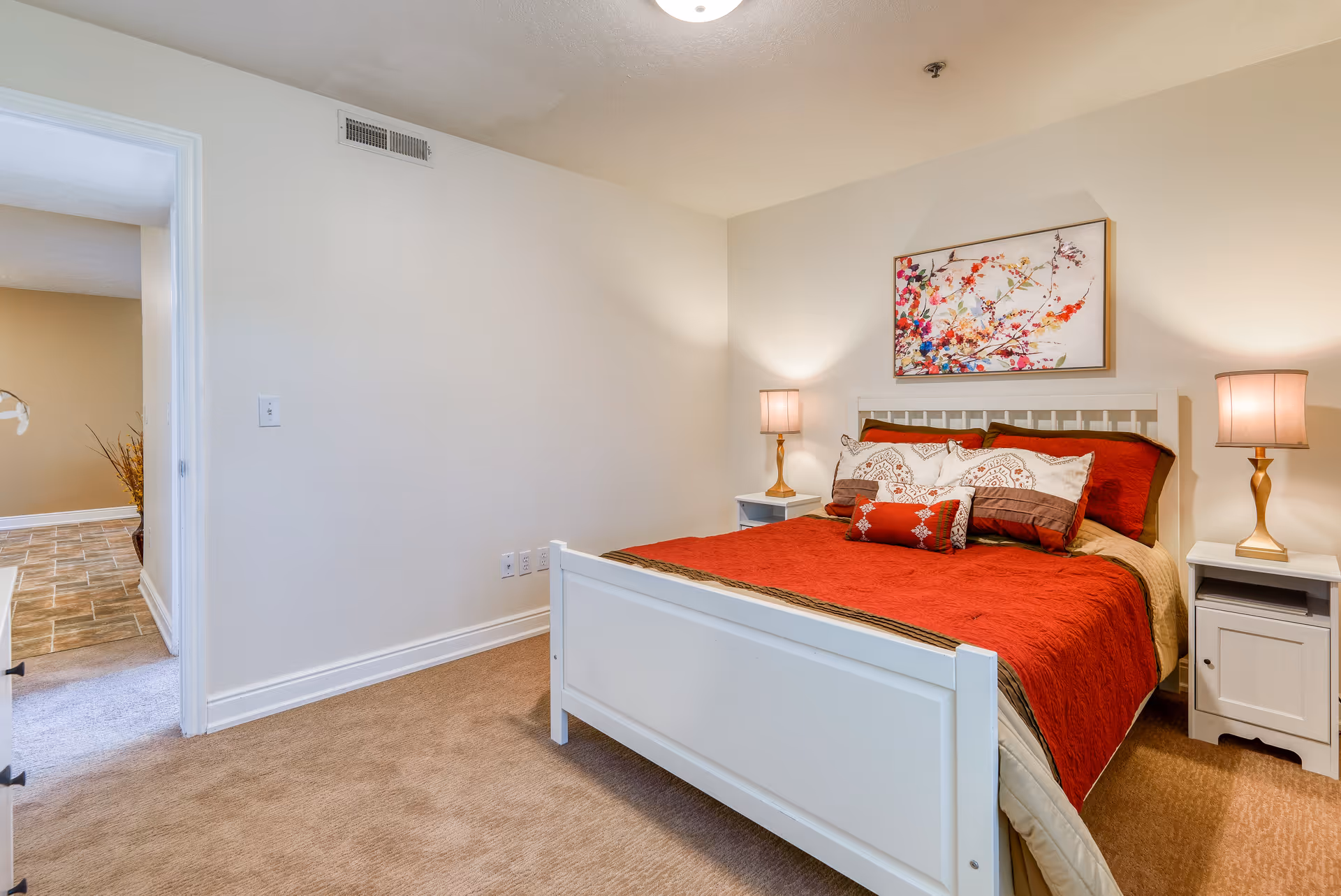 A tidy bedroom with a white bed dressed in red bedding, matching nightstands and lamps, and floral artwork above the headboard.