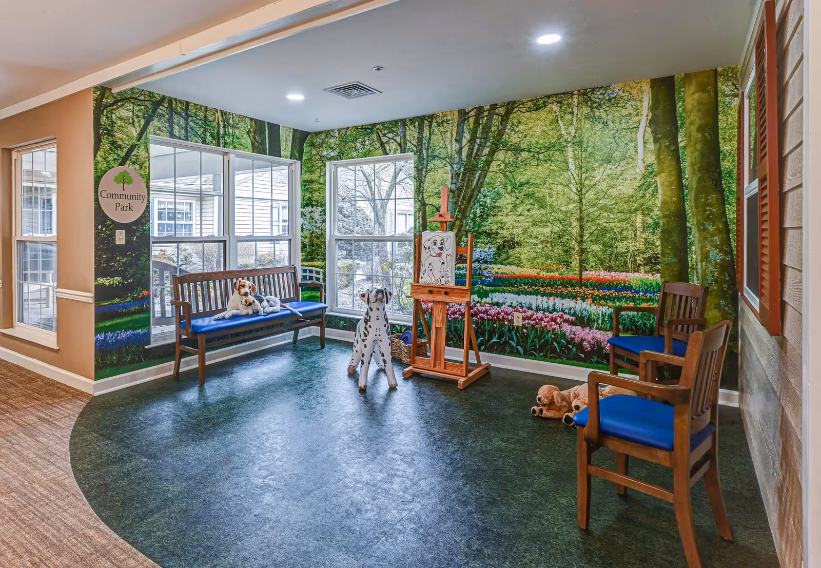 A cozy indoor sitting area with wooden chairs and a bench with blue cushions. The walls feature a large mural of a lush green forest and colorful flower garden, creating a park-like atmosphere. There are stuffed animal dogs placed on the bench, floor, and an easel with a drawing of a dog. A sign on the wall reads 'Community Park'.