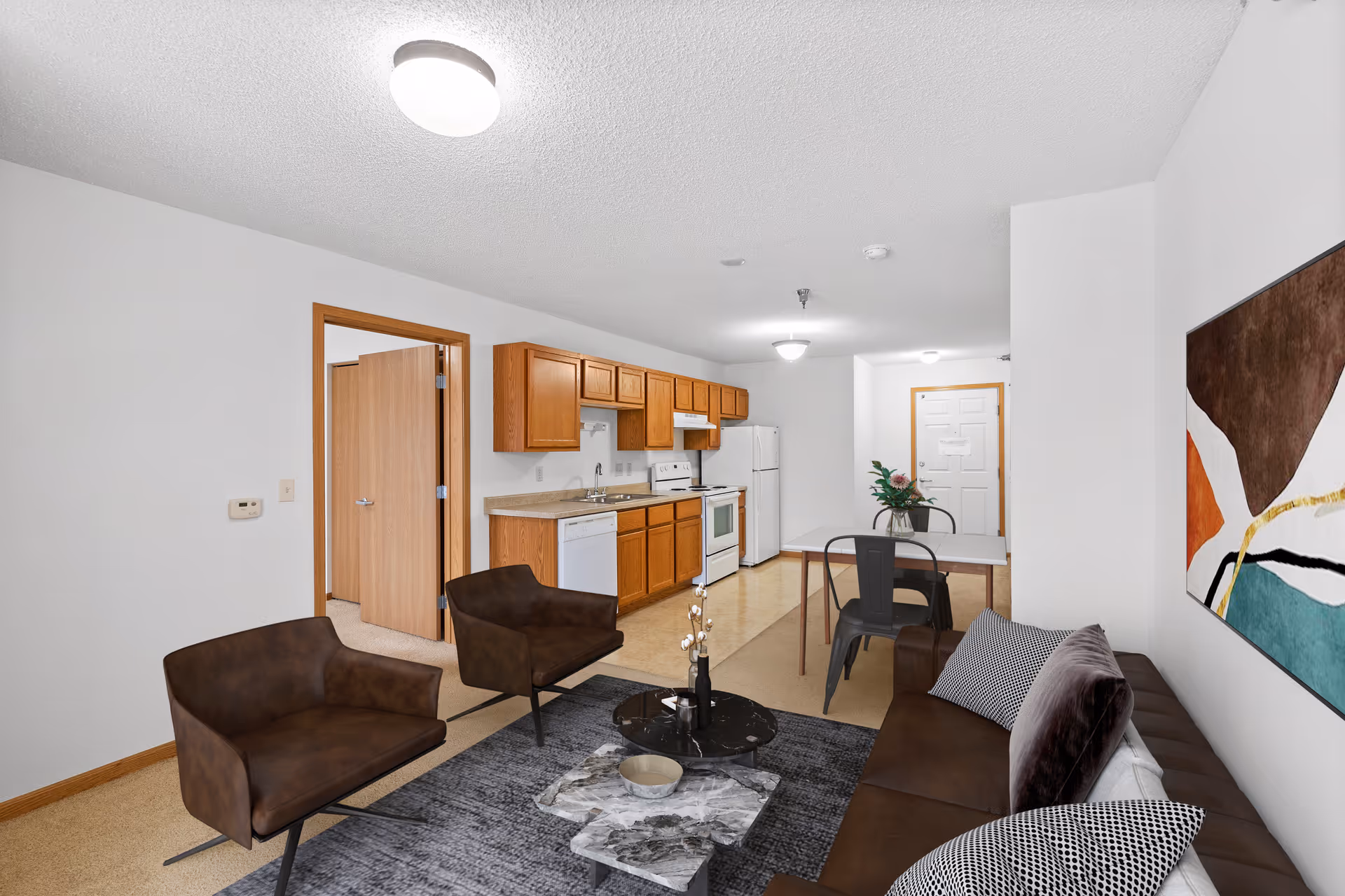 Interior view of a senior living apartment featuring a living area with a brown sofa, two brown armchairs, a coffee table, and a colorful abstract painting on the wall. The space opens into a kitchen with wooden cabinets, a white stove, refrigerator, and dishwasher. A dining table with two chairs and a vase with flowers is positioned near the kitchen. An open door leads to another room.
