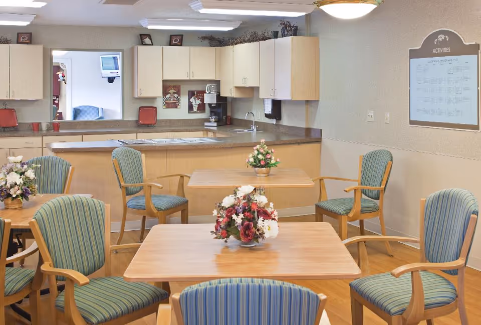 A communal dining area with wooden tables and striped cushioned chairs. Each table has a floral centerpiece. In the background, there is a kitchenette with light wood cabinets, a coffee maker, a sink, and a countertop. On the wall to the right, there is an activities board.
