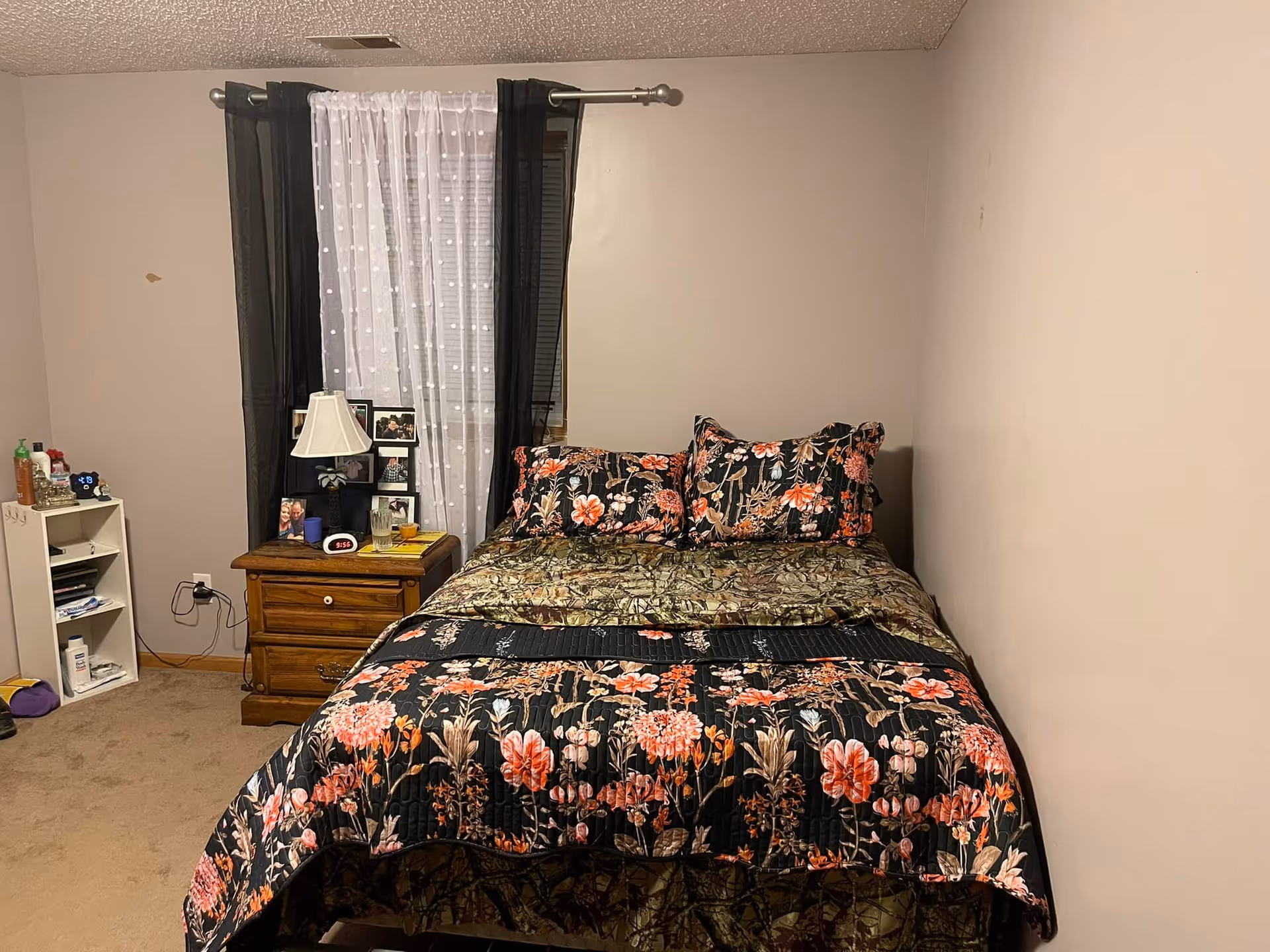 A bedroom with a floral quilted bed, wooden nightstand with a lamp and photos, and a curtained window.