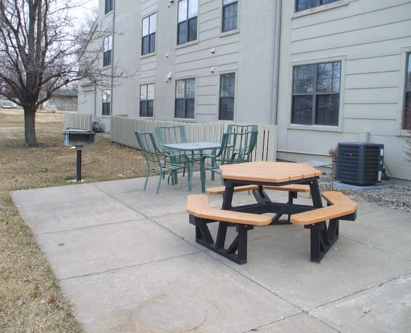 Outdoor patio area next to a building with a concrete walkway, featuring a hexagonal wooden picnic table with attached benches, a metal table with four metal chairs, a charcoal grill, and a leafless tree on a grassy lawn.