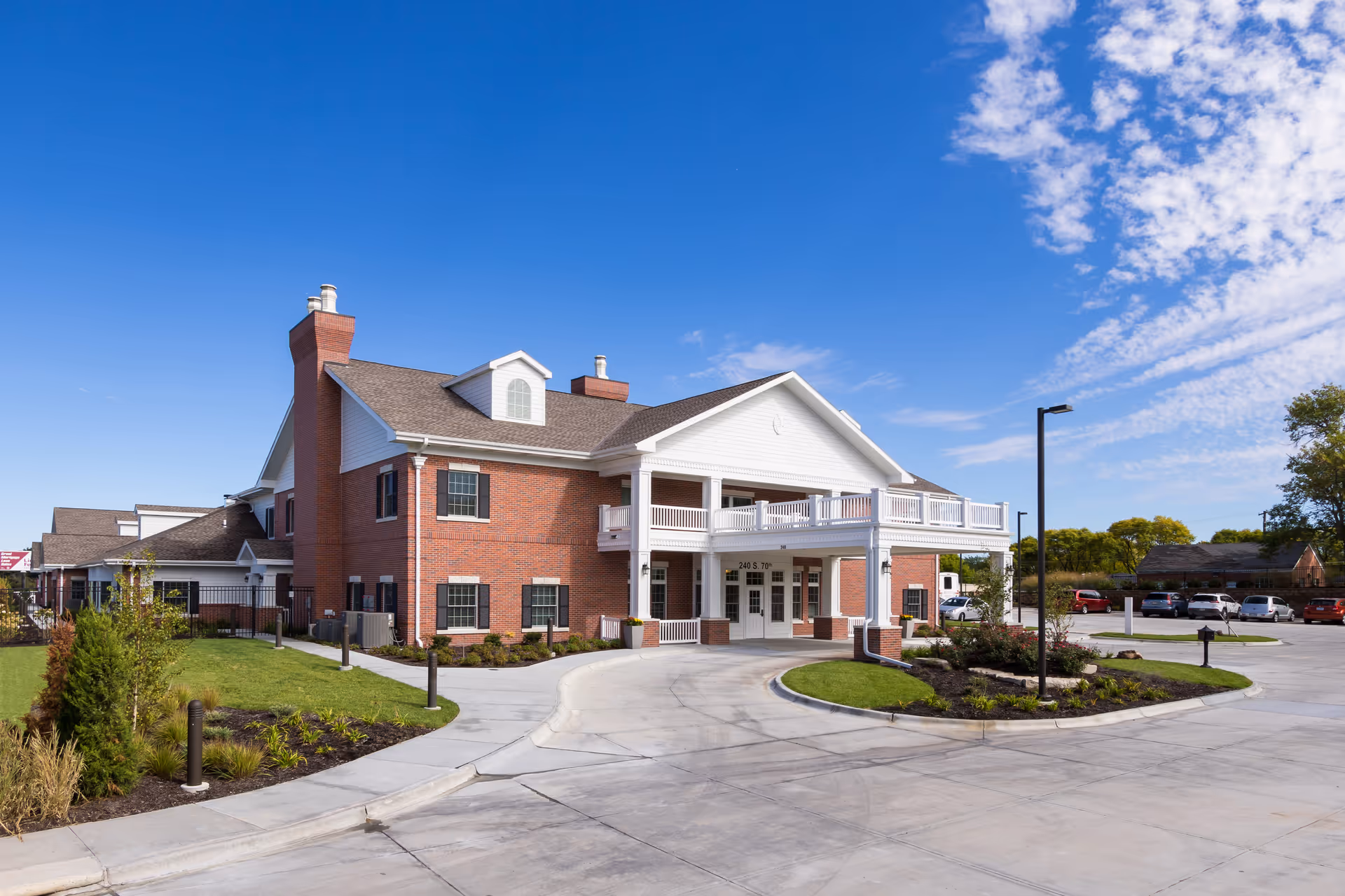 Exterior view of a two-story brick building with white trim and a covered entrance. The building has a driveway and landscaped areas with grass and shrubs. Several cars are parked in the parking lot under a clear blue sky.