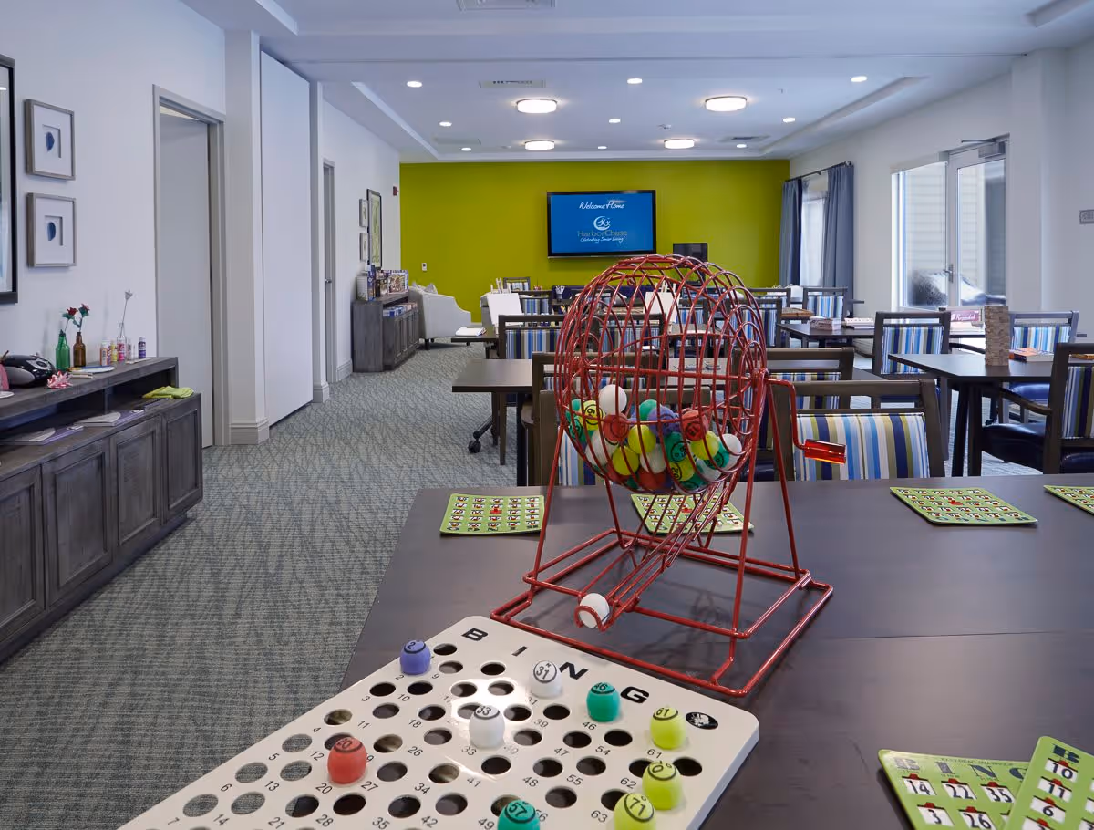 A bright and spacious common activity room with multiple tables and chairs arranged for group activities. In the foreground, there is a bingo game setup with a red bingo cage filled with colorful balls and a bingo card with markers. The back wall is painted green with a mounted TV displaying a welcome message for HarborChase Of Wilmington. The room has carpeted floors, framed artwork on the walls, and large windows with curtains allowing natural light.