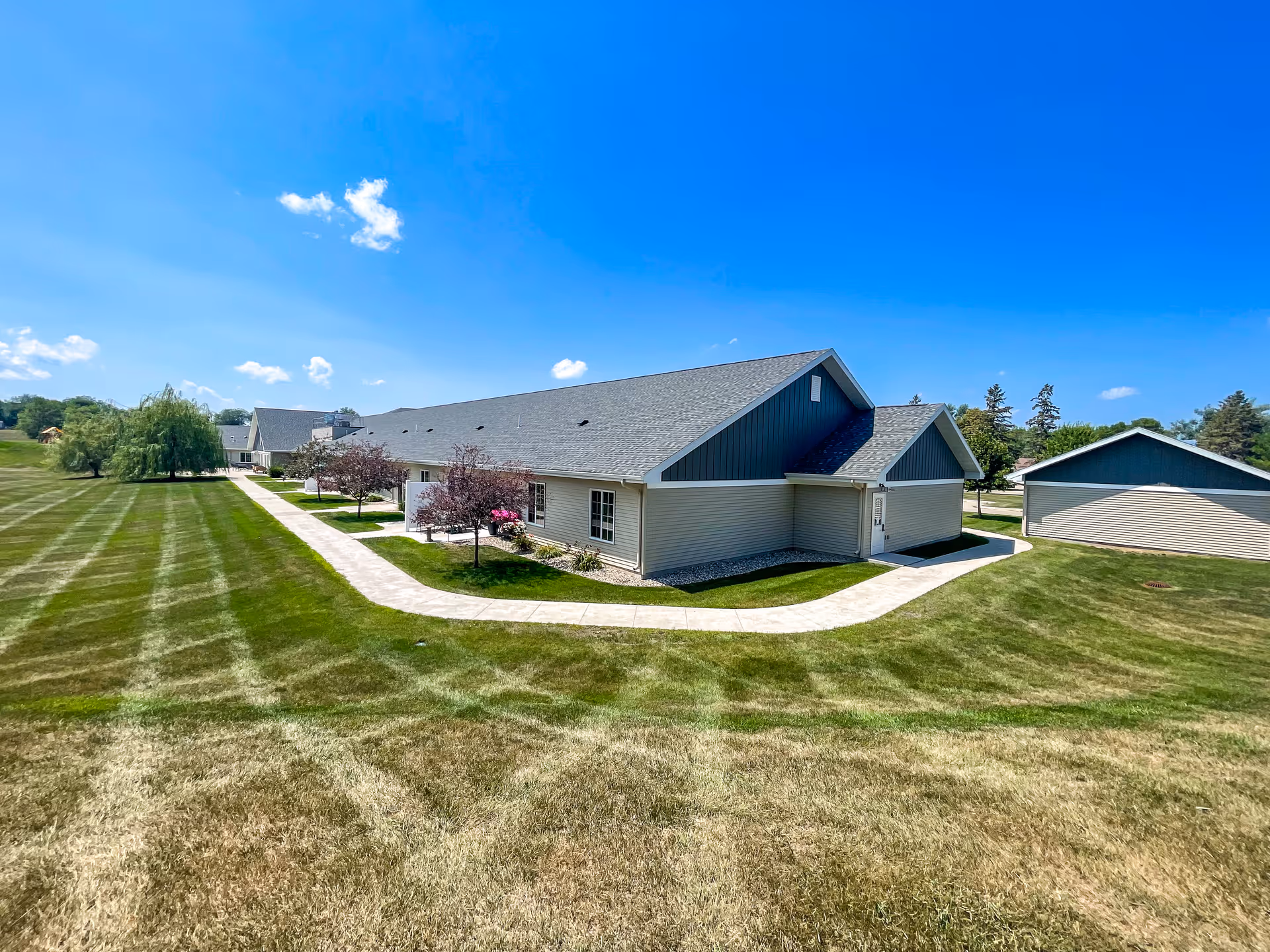 Exterior view of a single-story building with beige siding and a gray roof, surrounded by a well-maintained lawn and a paved walkway. The sky is clear and blue with a few scattered clouds. There are small trees and shrubs planted near the building.