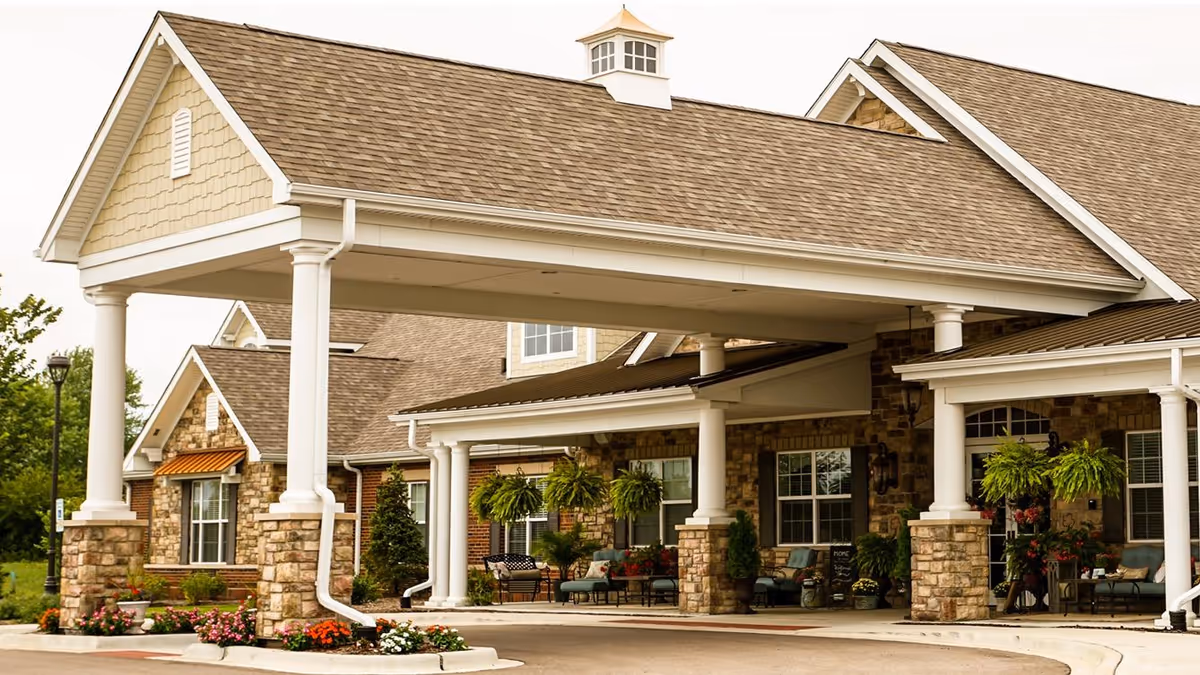 Entrance of a senior living facility with a covered drop-off area supported by white columns and stone bases. The building features a combination of stone and siding exterior with large windows and hanging plants. There are outdoor seating areas with chairs and small tables near the entrance.