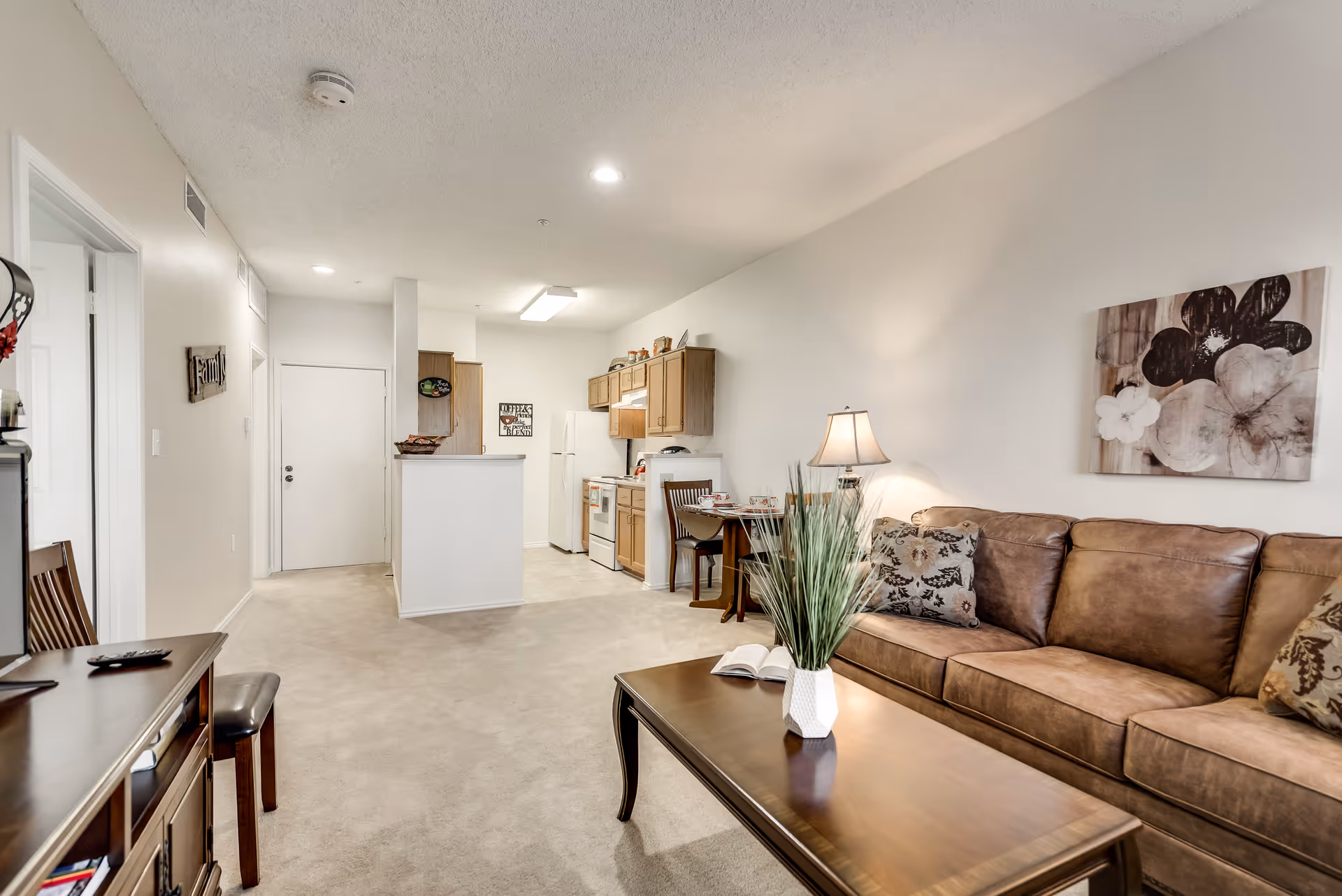 Interior view of a senior living apartment showing a living room with a brown leather sofa, a wooden coffee table with a white vase and green plant, a lamp on a side table, and a floral wall art. The background includes a small dining area with a table and chairs, and a kitchen with wooden cabinets, a white refrigerator, and a stove.
