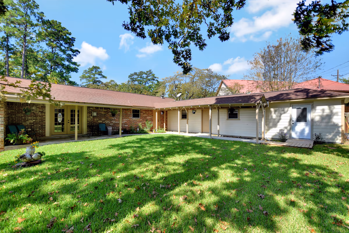 A sunny outdoor courtyard area of a single-story building with a covered walkway supported by columns. The building has a mix of brick and light-colored siding exterior walls, with several doors and windows. The courtyard features a well-maintained green lawn with some scattered leaves and shadows from nearby trees. The sky is blue with a few clouds, and tall trees surround the area.