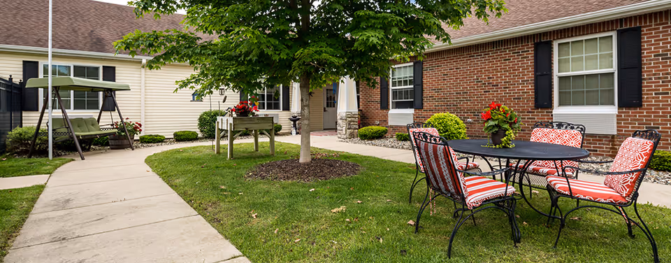 Outdoor courtyard area at Bickford of Battle Creek featuring a concrete walkway, a green cushioned swing, a black metal table with four chairs that have red and white patterned cushions, a tree in the center of a grassy area, and brick and siding building walls with windows in the background.