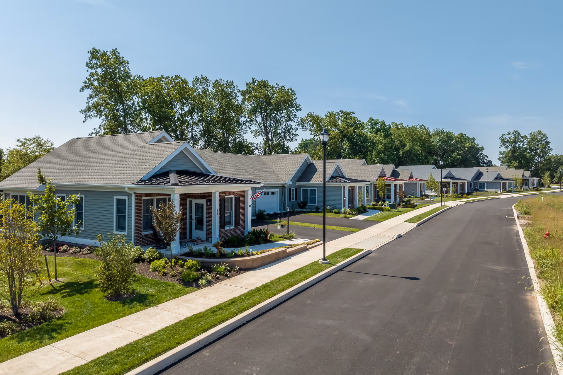 A row of single-story residential buildings with gray siding and brick accents along a paved road, surrounded by green lawns, small trees, and shrubs under a clear blue sky.