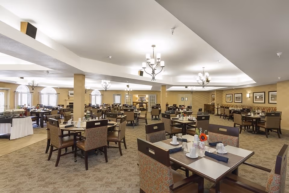 Spacious dining room with multiple tables and chairs arranged neatly. Each table is set with placemats, napkins, cups, and small flower vases. The room features beige walls, carpeted floors, and several chandeliers hanging from the ceiling. Large windows with arched tops allow natural light to enter the space.