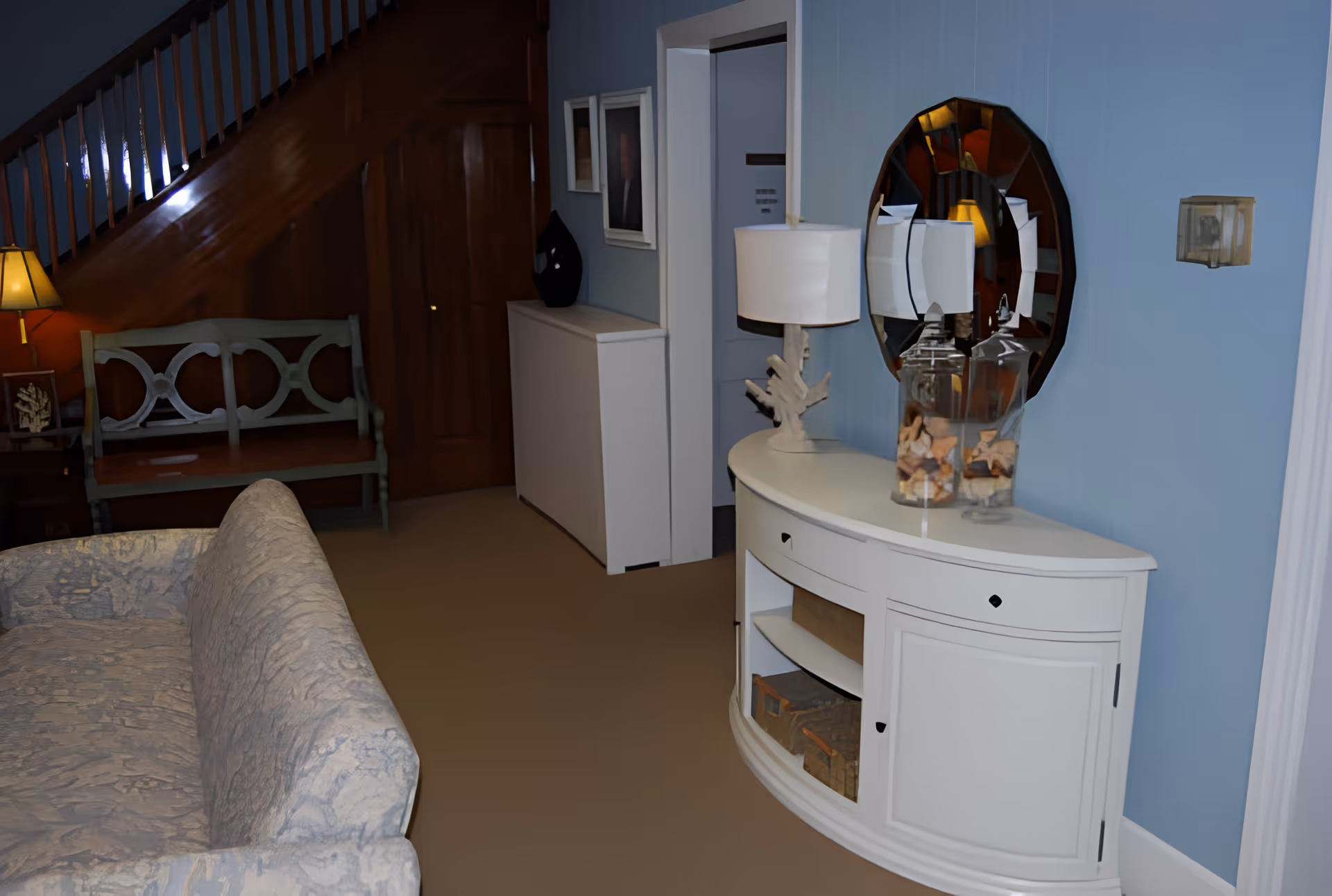 Cozy living room area with a patterned sofa, white console table topped with a lamp and mirror, and a wooden staircase and bench in the background.
