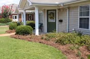Exterior view of a single-story residential building with a green front door, white columns, and windows. The building is surrounded by neatly trimmed bushes, mulch, and a well-maintained lawn. A concrete walkway leads to the entrance.
