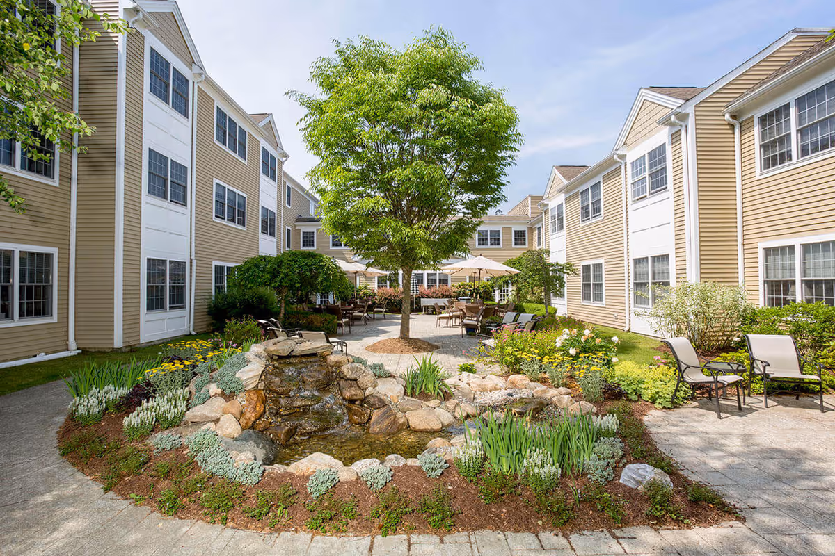 Outdoor courtyard area at Benchmark at Ridgefield Crossings featuring a small rock waterfall pond surrounded by plants and flowers, a central tree, patio seating with tables and umbrellas, and beige multi-story buildings with white trim in the background under a partly cloudy sky.