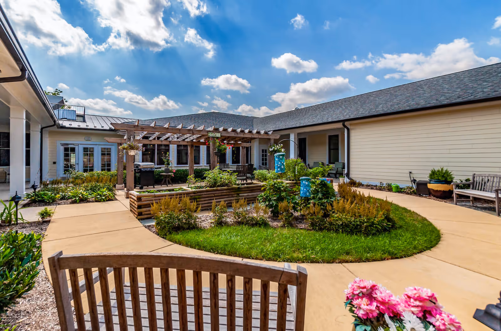 Outdoor courtyard area at Chantilly Heights Memory Care featuring a wooden pergola with hanging plants, garden beds with various plants and flowers, a paved walkway, wooden benches, and a bright blue sky with scattered clouds.