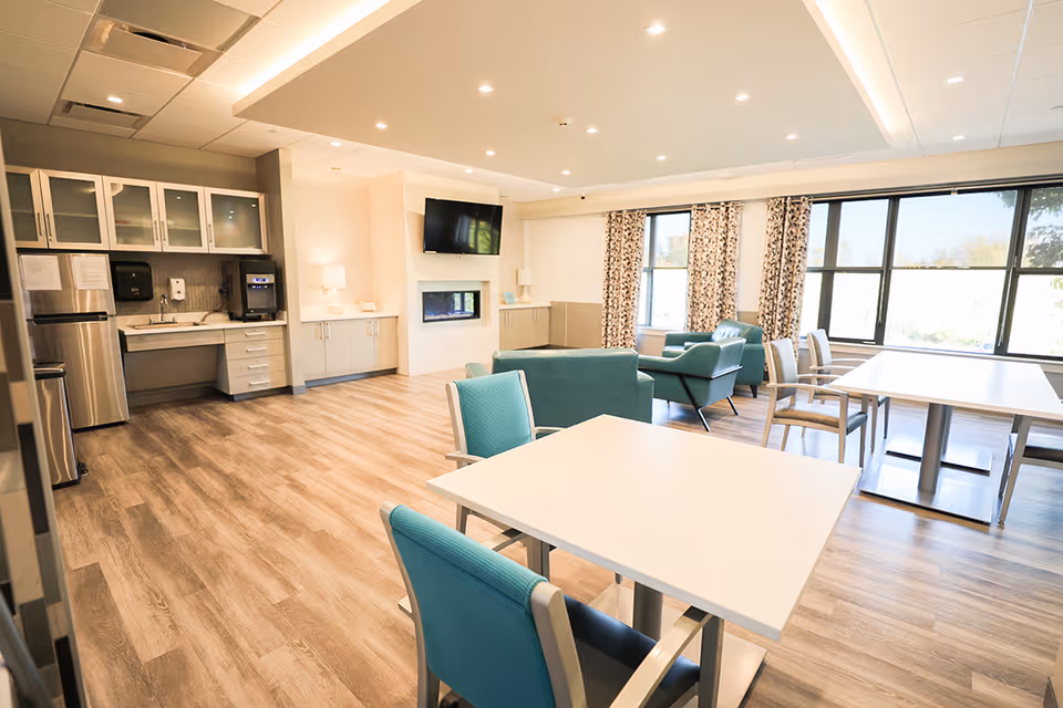 A bright, spacious common area in a nursing and rehabilitation center featuring a kitchenette with stainless steel appliances, white cabinets, and a sink on the left. The room has wood flooring, several tables with teal and beige chairs, a seating area with teal armchairs, large windows with patterned curtains, a wall-mounted TV above a modern fireplace, and soft lighting from ceiling fixtures and lamps.