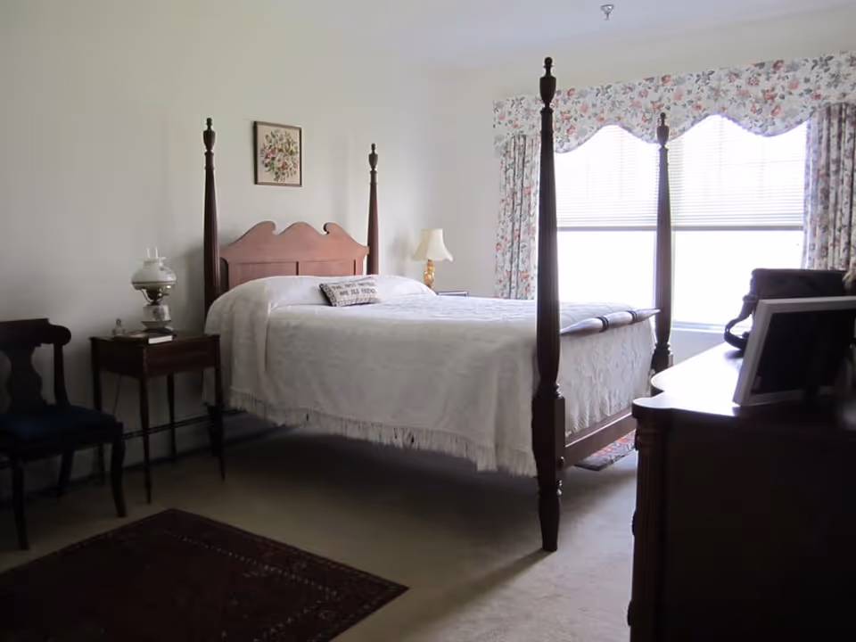 A bedroom with a wooden four-poster bed covered with a white bedspread. There is a small wooden side table with a lamp and a chair next to it on the left side of the bed. A window with floral curtains allows natural light into the room. A dresser with a framed photo is visible on the right side.