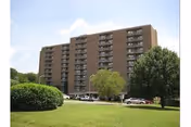Front view of a multi-story brick residential building with balconies, parked cars, and a grassy lawn with trees.